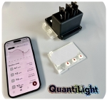 Zu sehen ist eine kleine schwarze Box mit nach oben ragenden Anschlüssen, eine durchsichtige Plastikkartusche und ein Mobiltelefon mit geöffneter App und den Testergebnissen.